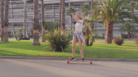 Beautiful young woman rides fast skateboard through the Park past palm trees and modern building Sunny day. Tracking shot. 60 fps.