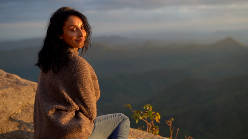 The sunset lights up the face of a youg woman sitting on a cliff overlooking the mountain range below - slow motion