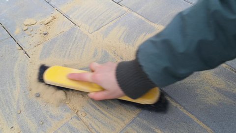 Woman's hand wipes seams between new paving tiles with a brush-whisk. The final stage of laying paving slabs - filling gaps with sand