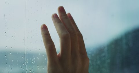 Close up of a young woman hand is touching a glass with water drops while outside is raining.