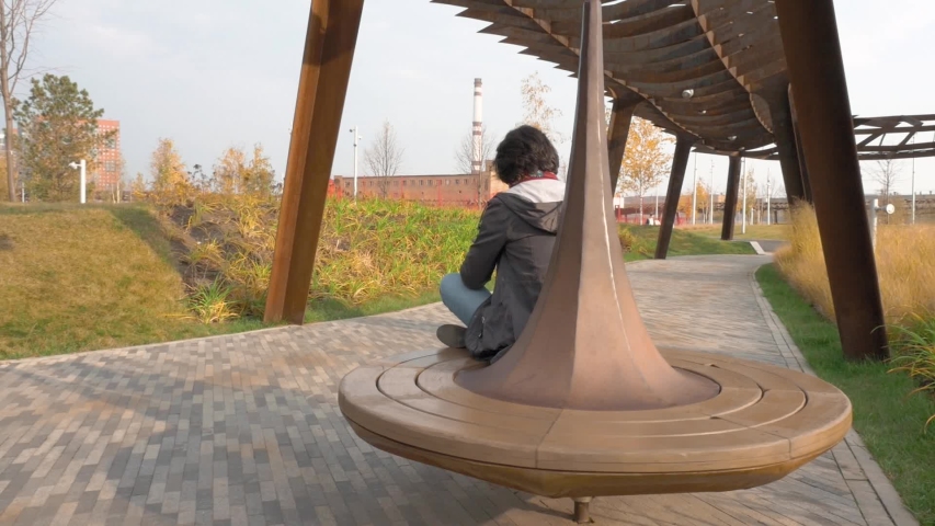 Caucasian middle aged woman brunette black jacket Burgundy scarf blue jeans riding on carousel in Park Sitting cross legged Holding smartphone Clicks in background stone buildings Around lawn