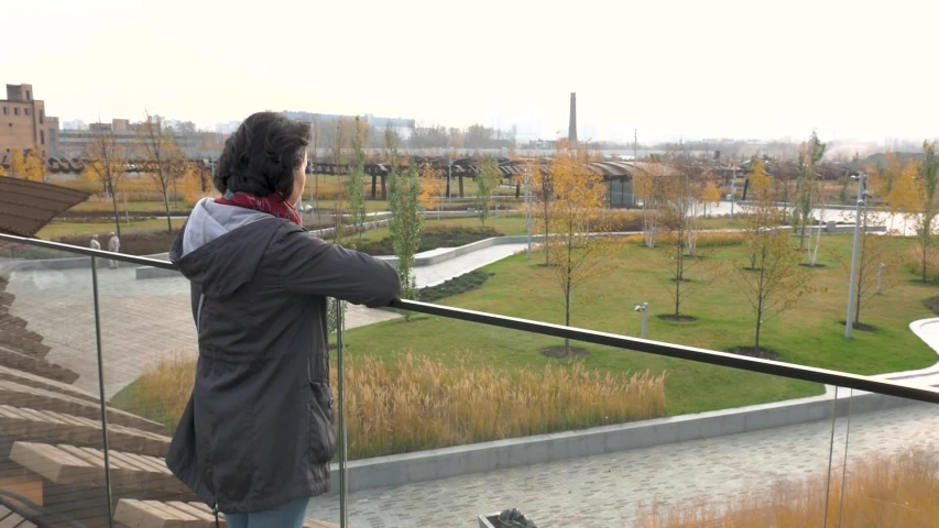 A small parkland Caucasian brunette woman in a black jacket stands at the glass parapet looking ahead. The camera moves on it then shifts the angle to the territory of the Park Autumn day | Shutterstock HD Video #1041910978