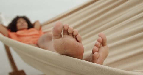 Front view of an African American woman on a beach by the sea, lying in a hammock asleep, feet facing camera, slow motion