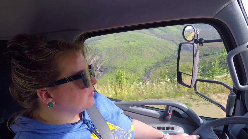 Static shot, of a girl with sun shades, driving a van, on a sand road, up a steep hill, on the Taylor Highway, on a partly sunny day, in Alaska, USA