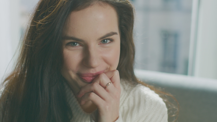 Portrait of Beautiful Young Brunette Smiling Charmingly while Sitting on a Sofa, Brushes Her Lush Hair Away with a Flirtatious Gesture. Shy Girl Wearing Oversized White Knitted Sweater Relaxes  Royalty-Free Stock Footage #1041916387