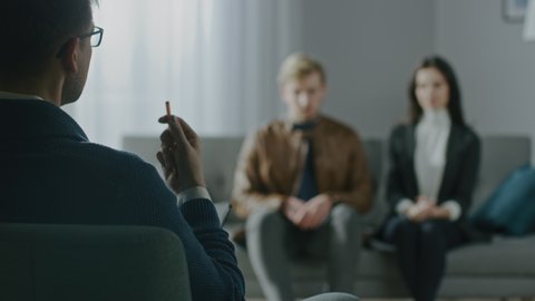 Unfocused Couple on a Counseling Session with Psychotherapist. Foreground Focus on Back of the Therapist Taking Notes and Talking: People Sitting on the Analyst Couch, Discussing Relationship 