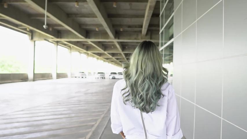 Asian Teenager girl turns around and looks at camera. Beautiful woman has long Green curl hair. Attractive fashion model is walking, shaking and flying curly green coloring hair. Slow Motion Gimbal