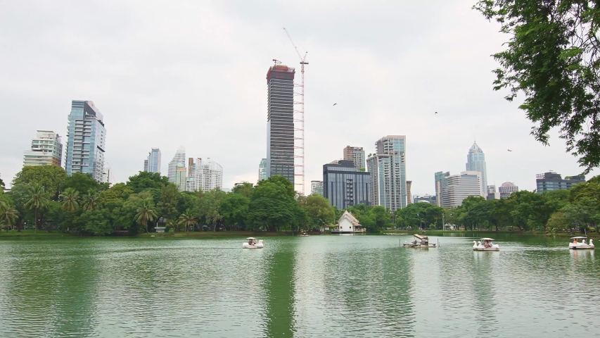 Central park Lumpini in Bangkok city, Thailand. Panoramic view of cityscape with skyscrapers and modern futuristic buildings. View of lake in the park with swan boat. Reflection in the water.