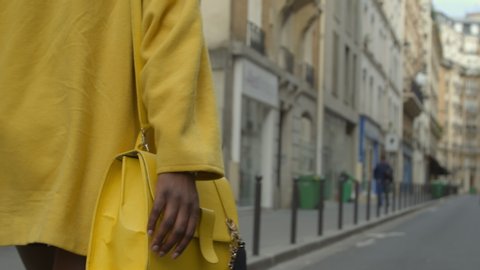 Camera follows a glamourous black woman wearing a bright yellow jacket and matching handbag walking down a shopping district in France. Handheld, close-up, 4K, CU, slow motion, people, person, single.