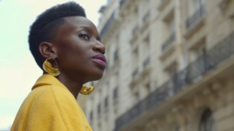 Beautiful African woman looking out at city confidently while she waits on transportation. Portrait of gorgeous fashion model looking boldly outward as she travels in Paris, France. 4K, CU, slow-mo.