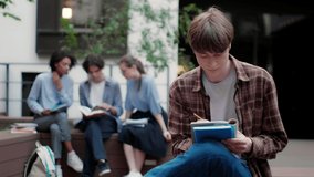 Medium shot of casual student boy thoughtfully studying alone with group of students on background in university campus outdoor