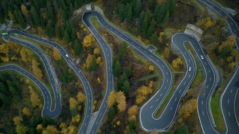 Drone zooms out from beautiful mountain road on high altitude alpine pass. Winding turns and switchbacks in orange autumn forest, cars and buses drive carefully