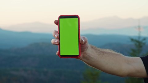 Hand of man showing mock-up modern smartphone typing on chroma green screen application on beautiful background. Blurred view of scenic mountains and blue sky.