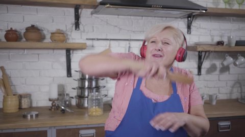 Close-up of positive senior Caucasian woman dancing and emotionally gesturing. Funny old woman in headphones singing and dancing at the kitchen. Mature cheerful retiree enjoying life after retirement.
