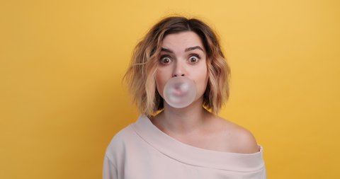 Portrait happy smile woman inflates bubble from chewing gum and bursts it in pink sweater slow motion on an isolated yellow background. Teen girl happiness. Monotone. Emotions of people. Lifestyle