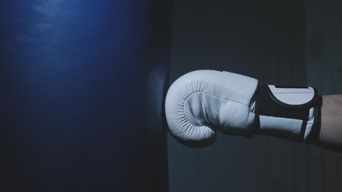 Close-up of boxer hits punching bag in dark gym in slow motion. Young man training indoors. Strong athlete in gym. Sport concept. Sportsman boxing in smoky studio