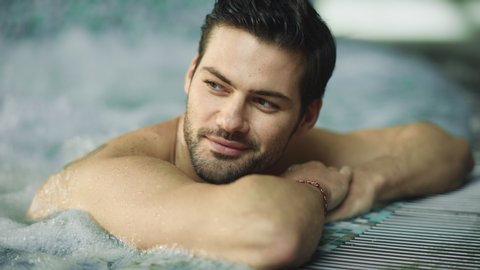 Portrait of sexy man enjoying in whirlpool bath in slow motion. Closeup handsome man relaxing in pool at wellness hotel. Attractive man having hydrotherapy in jacuzzi spa indoor.