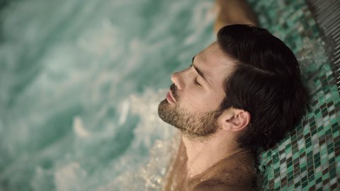 Close up of sexy man enjoying jacuzzi spa in slow motion. Portrait of handsome guy relaxing in pool at wellness hotel. Attractive man having hydro massage in jacuzzi indoor.