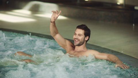 Attractive man resting whirlpool bath indoor. Happy man relaxing in pool spa at wellness center. Smiling guy saying hi at spa hotel. Sexy businessman enjoying rest at spa hotel.