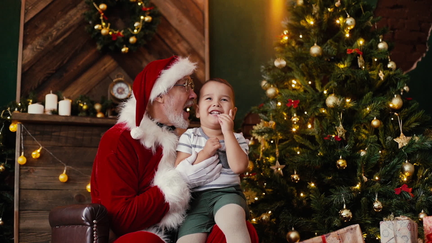 Little cute boy making wish sitting on Santa Claus laps, next to the Christmas tree in room with festive interior. Shooting in slow motion.