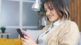 Young Woman Seat on Couch in Modern Living Room and Using Smartphone for Watching Video or Social Network. Lifestyle Concept. Happy Woman Relaxing at Home.