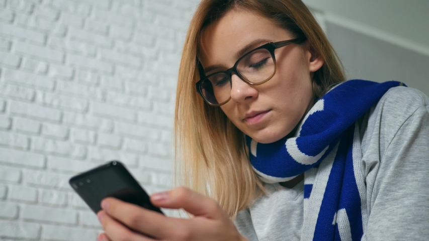 A young Caucasian woman stares sports game using the app on a smartphone while sitting on a couch in the house at night. - Powered by Shutterstock - Get 15% off with code: PIKWIZARD15