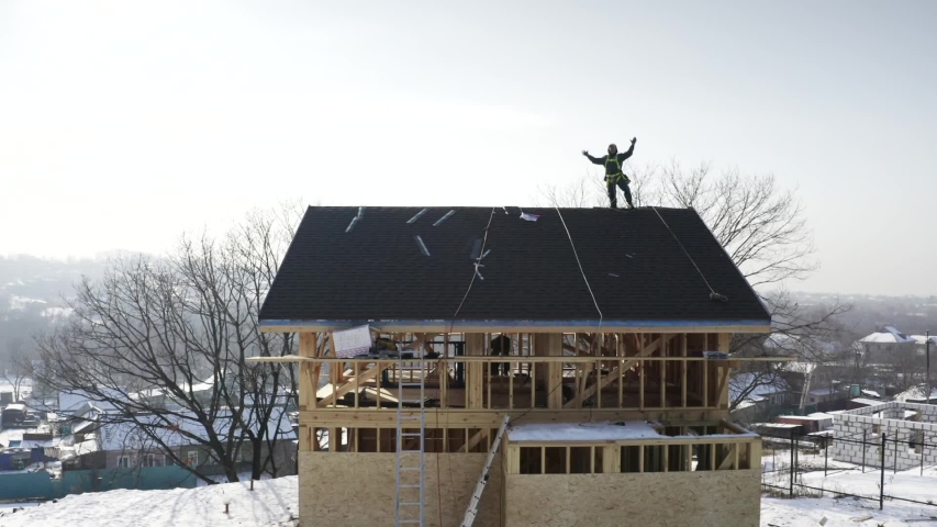 Aerial panoramic view of residential house under constuction and tiler in safety harness on the roof top waving into the camera. Bright sunny day