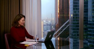 a woman in a Burgundy dress sits at a round table near a large window. she communicates through a video link on a laptop and makes recordings. behind the window you can see high-rise city buildings
