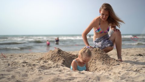 mother in bikini bury her little blond daughter girl in the beach sand. camera movement closer shot with gimbal.