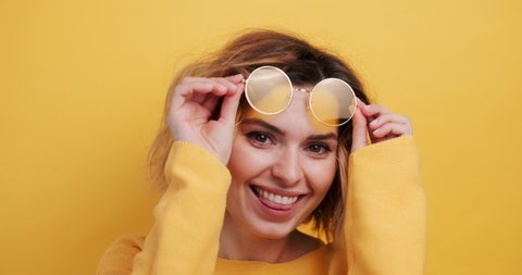 Portrait of happy young woman correcting yellow glasses on her forehead coquettishly showing tongue to camera smiling on an isolated yellow background. Monotone. Emotions of people. Copy space