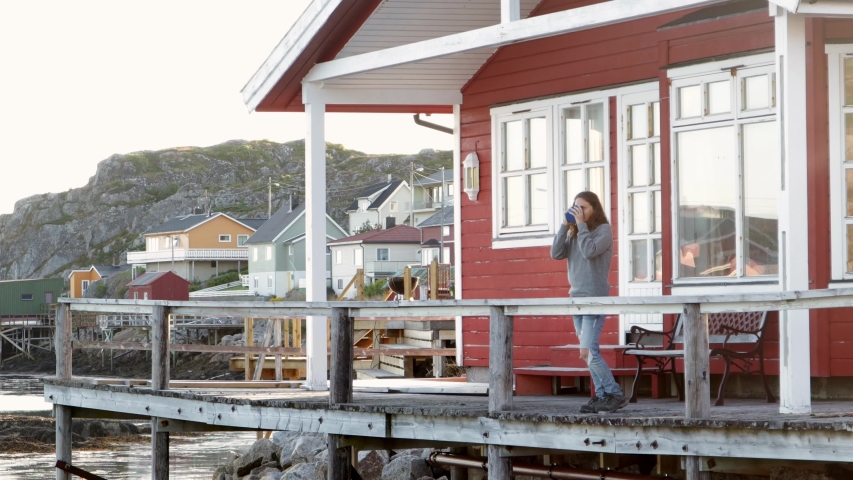 A young woman standing at the fishing huts terrace at sunset time and enjoys the view. Norwegian traditional rorbu at the beautiful Fjord.
