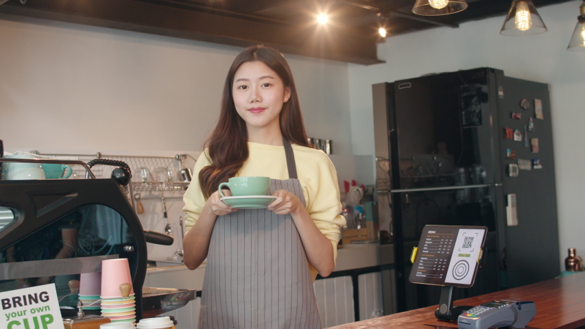 Portrait young Asian lady barista waitress holding coffee cup feeling happy at urban cafe. Asia small business owner girl in apron relax toothy smile looking to camera stand at counter in coffee shop. Royalty-Free Stock Footage #1042013854