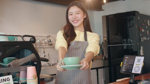 Portrait young Asian lady barista waitress holding coffee cup feeling happy at urban cafe. Asia small business owner girl in apron relax toothy smile looking to camera stand at counter in coffee shop.