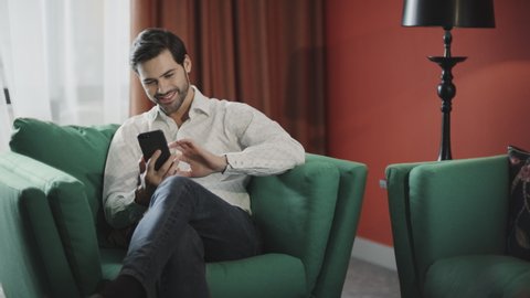Young businessman looking mobile phone at luxury hotel. Business man relaxing phone at hotel room. Smiling guy scrolling smartphone in modern apartment.