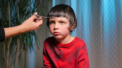 Hair cutting at home. Epidemic of coronavirus infection COVID-19. Hairdressers are closed.A serious child on a haircut by a hairdresser. A boy in red clothes is scissored by cutting bangs on his head.