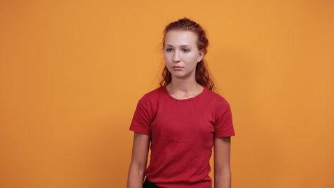 Disappointed young lady in red shirt keeping hand up, pointing aside isolated on orange background in studio. People sincere emotions, health concept.