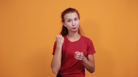 Pretty young lady in red shirt keeping fists up, pointing directly, looking serious isolated on orange background in studio. People sincere emotions, health concept.