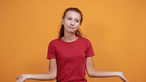 Disappointed young woman in red shirt isolated on orange background in studio spreads arms, raising shoulders. People sincere emotions, health concept.