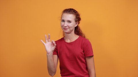 Pretty young lady in red shirt showing okay gesture, smiling, laughting isolated on orange background in studio. People sincere emotions, health concept.