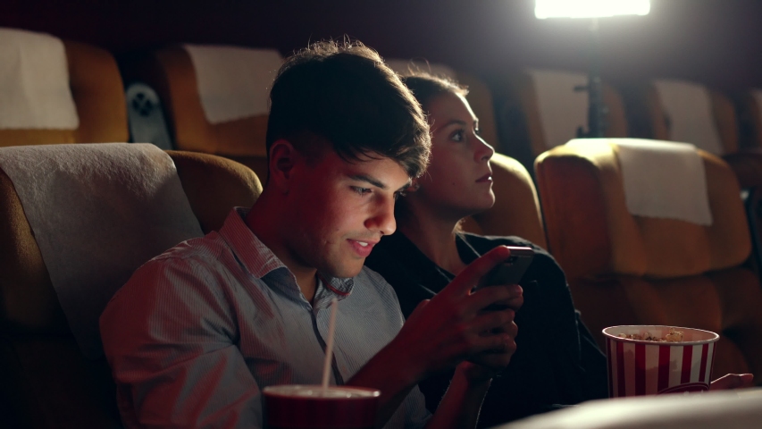 A man is playing a mobile phone game. He is happy because he wins the game while his girlfriend is watching a movie next to him.