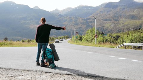Young woman hitchhiker standing on the road with a view to the mountains. Hitchhiking at Lofoten Islands in Norway.