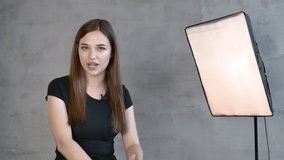 Caucasian Girl in a black t shirt long straight hair brown haired on the background of a gray wall and a lamp emotionally speaks to the camera takes his hand to the side bows his head to the side