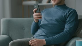 Portrait of Handsome Young Man Using Smartphone, While Sitting on a Chair in His Cozy Living Room. Creative Freelancer Relaxes at Home, Surfs Internet, Uses Social Media, Watches Videos. Portrait 