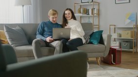 Cute Young Couple Use Laptop Computer, while Sitting on the Couch in the Cozy Apartment. Couple Surfing Web, Shopping on Internet, Using Social Media, Watching Videos and Streaming Content