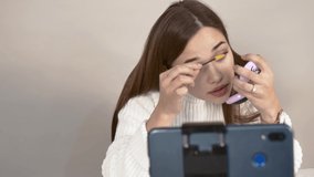 Grey background Caucasian girl video blogger white jumper straight hair brown haired sits in front of phone camera Tassel paints her eyebrows Close up