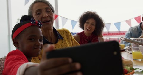 Front view close up of a young African American girl sitting at the dinner table at home taking a selfie with her grandmother, with her mother in the background during a family meal, slow motion