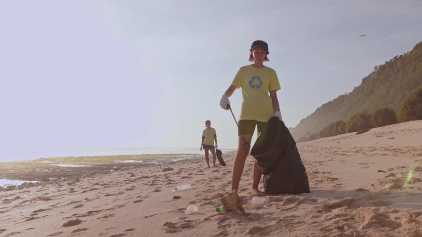 A female volunteer wearing a green t-shirt with recycling sign print, tidying up rubbish with a trash grabber stick and put in garbage bags, cleaning area on the white sand beach, eco-friendly Royalty-Free Stock Footage #1042070533