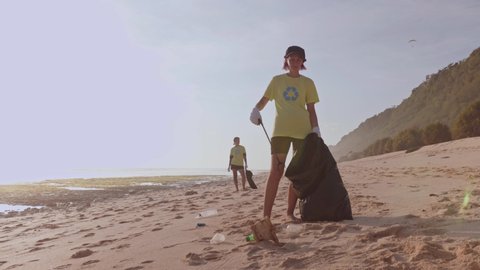A female volunteer wearing a green t-shirt with recycling sign print, tidying up rubbish with a trash grabber stick and put in garbage bags, cleaning area on the white sand beach, eco-friendly
