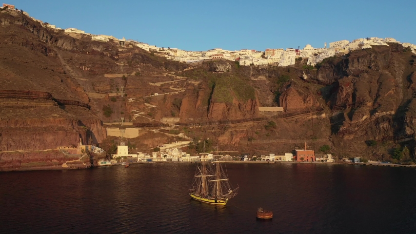 Aerial drone video of wooden sail boat docked near old port of Santorini island in deep blue sea just below village of Fira at sunset, Cyclades, Greece