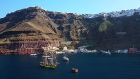 Aerial drone video of wooden sail boat docked near old port of Santorini island in deep blue sea just below village of Fira at sunset, Cyclades, Greece